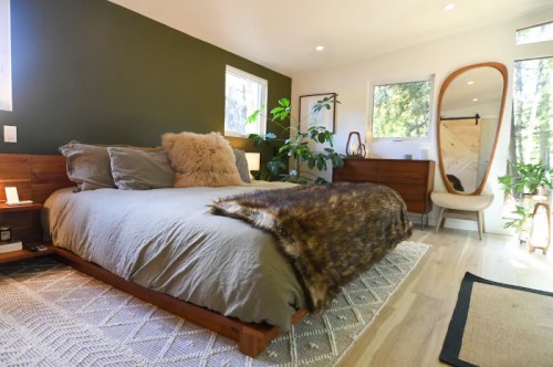 Cozy bedroom with a wooden bed, plush bedding, a large mirror, and greenery, featuring warm, natural tones.