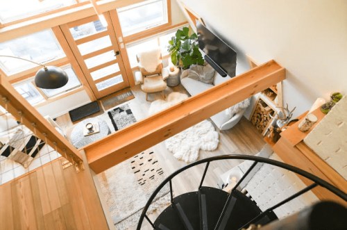 A cozy, modern living room with a spiral staircase, large windows, and a plant, featuring soft furnishings and warm wood accents.