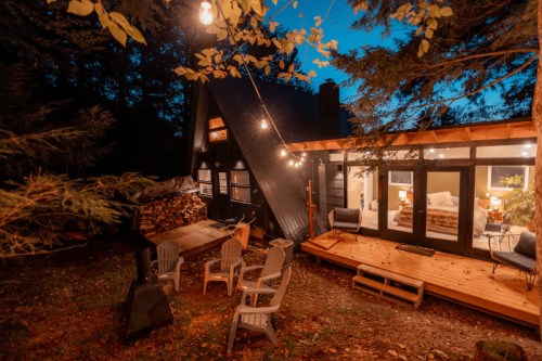 Cozy cabin at night, featuring string lights, a wooden deck, and a fire pit surrounded by chairs in a wooded setting.