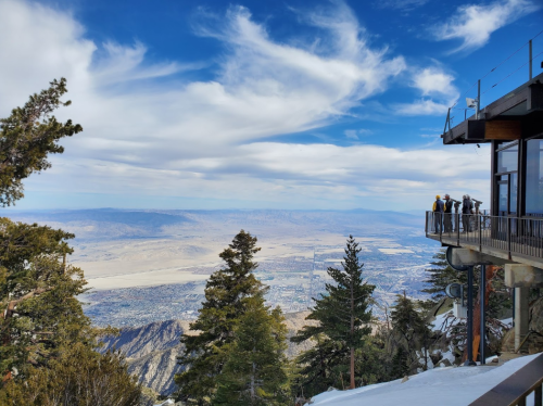 A scenic view from a mountain overlook, featuring trees, a building, and a vast valley under a blue sky with clouds.
