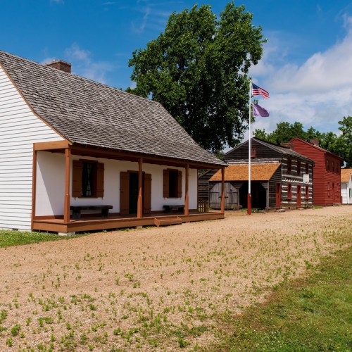A historic village scene featuring wooden buildings, a gravel path, and flags under a blue sky with scattered clouds.