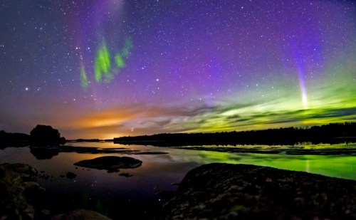 A vibrant night sky with green and purple auroras reflecting on a calm river, surrounded by dark silhouettes of trees.