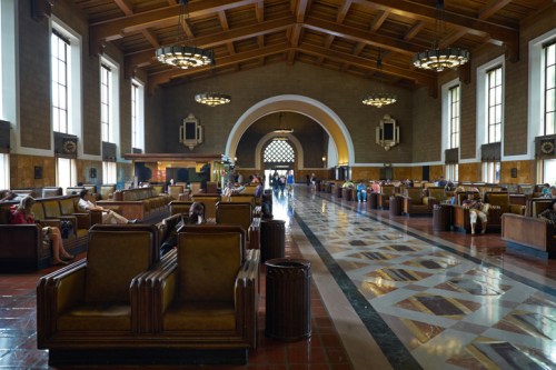 Interior of a spacious train station with wooden beams, large windows, and seating areas filled with people.