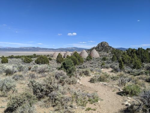 A desert landscape featuring several conical stone structures surrounded by shrubs and mountains in the background.
