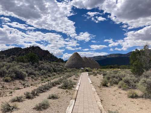 A pathway leads through a desert landscape to large, conical stone structures under a cloudy sky.