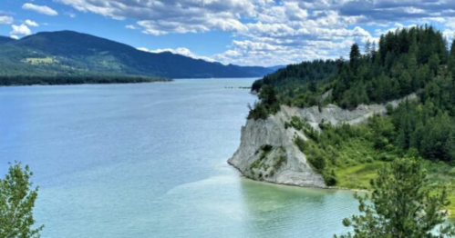 A serene lake surrounded by lush green hills and rocky cliffs under a blue sky with fluffy clouds.