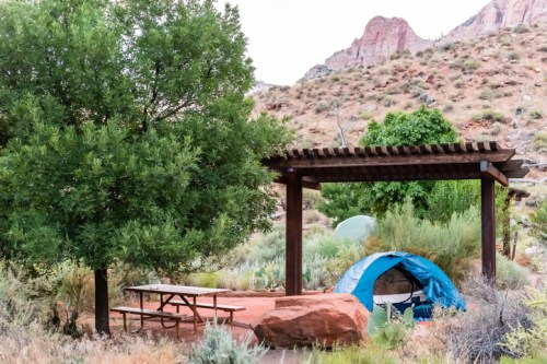 A blue tent and picnic table under a wooden pergola, surrounded by shrubs and rocky hills.