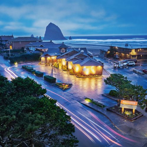 A coastal scene at dusk featuring illuminated buildings, a winding road, and a prominent rock formation in the ocean.