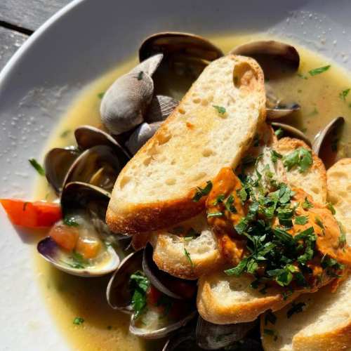 A bowl of clams in broth topped with toasted bread and a dollop of sauce, garnished with fresh herbs and diced tomatoes.