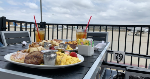 A breakfast spread with pancakes, eggs, sausage, and drinks on a patio overlooking an airport runway.