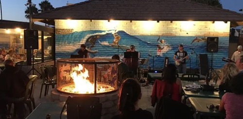 A lively outdoor scene with a band performing near a fire pit, surrounded by diners and a colorful mural of waves.