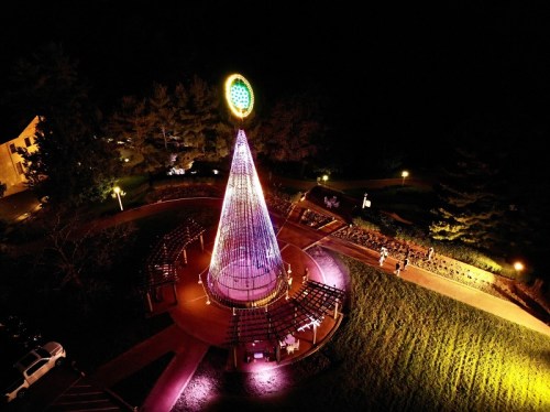A brightly lit Christmas tree made of lights stands in a park at night, surrounded by benches and trees.