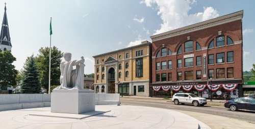 A statue stands in a plaza with buildings and a church in the background, decorated with red, white, and blue bunting.