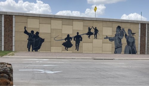 Silhouettes of dancers and performers on a mural against a beige wall, with a blue sky and clouds above.