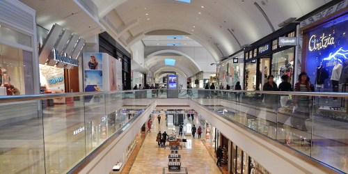 A spacious shopping mall interior with stores on both sides and shoppers walking along the central walkway.