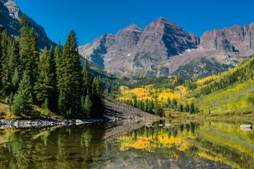 A serene mountain landscape with vibrant autumn trees reflecting in a calm lake under a clear blue sky in Colorado