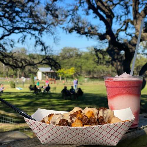 A tray of crispy food with sauce beside a colorful drink, set against a sunny park with people in the background.