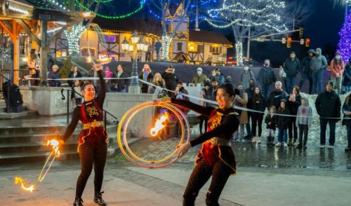 Two performers twirl flaming hula hoops in front of a festive crowd, with holiday lights in the background.