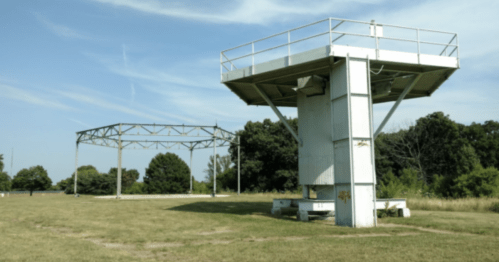 Two large, industrial structures stand in a grassy field under a clear blue sky, surrounded by trees.