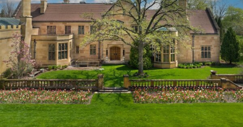 A large stone house with a manicured lawn and flower beds, surrounded by trees and greenery on a sunny day.