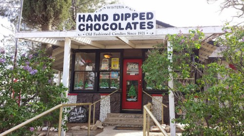 A charming storefront of "Hand Dipped Chocolates," featuring a sign and surrounded by greenery and flowers.