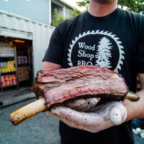 A person in a black shirt holds a large, smoked beef rib with a visible pink smoke ring, wearing gloves.