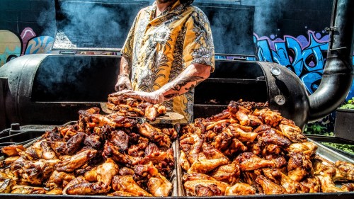 A person in a colorful shirt prepares grilled chicken wings on a large barbecue, surrounded by vibrant graffiti.