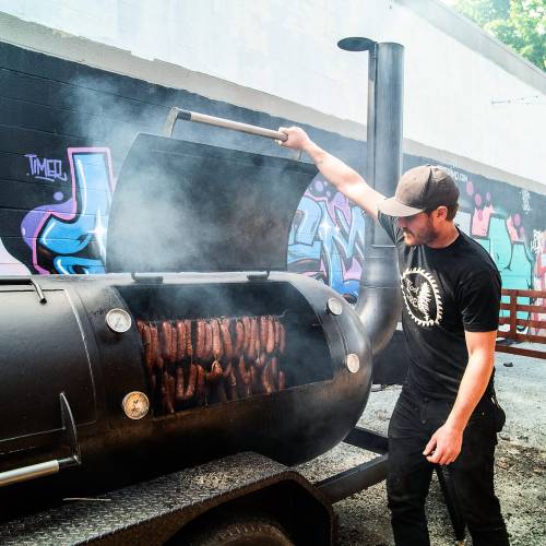 A person lifts the lid of a smoker filled with sausages, surrounded by colorful graffiti on a wall. Smoke billows out.