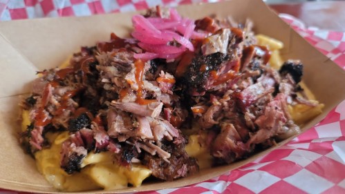 A close-up of a dish featuring pulled pork over creamy macaroni and cheese, topped with pink pickled onions and barbecue sauce.