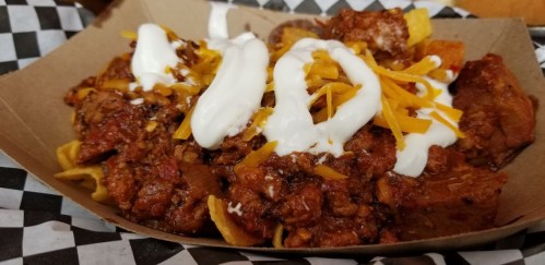 A serving of nachos topped with chili, shredded cheese, and sour cream, served in a brown paper tray on a checkered background.