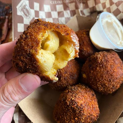 A hand holding a crispy, golden-brown mac and cheese ball, revealing creamy pasta inside, with a dipping sauce on the side.