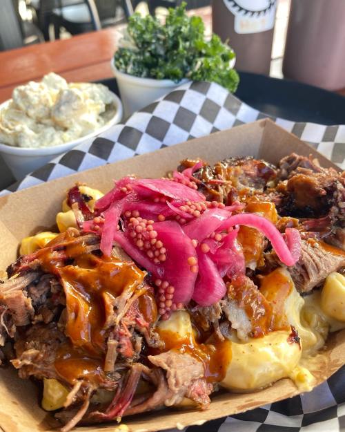 A close-up of a BBQ dish with pulled meat, mac and cheese, pickled onions, and mustard seeds, with sides of kale and potato salad.