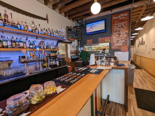 Interior of a BBQ restaurant featuring a bar with various drinks, a TV, and a staff member preparing food.