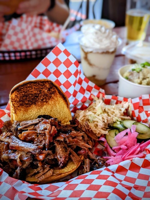 A sandwich filled with pulled meat on a checkered plate, accompanied by pickled vegetables and a dessert in the background.