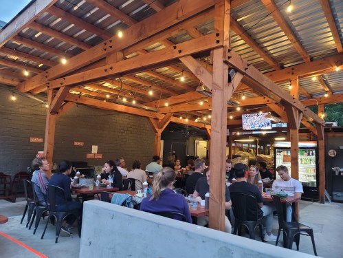 Outdoor dining area with wooden beams, string lights, and patrons enjoying meals at tables. A TV is visible in the background.