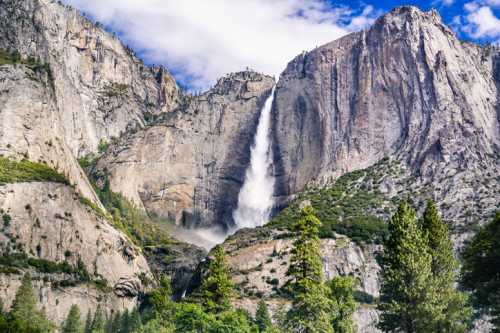 A majestic waterfall cascades down a rocky cliff, surrounded by lush greenery and towering mountains under a blue sky.