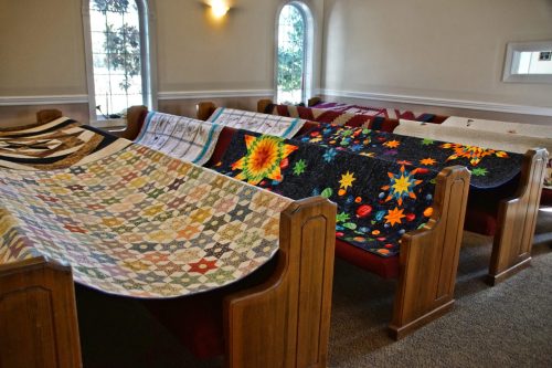 Colorful quilts displayed on pews in a church, showcasing various patterns and designs. Natural light filters in.