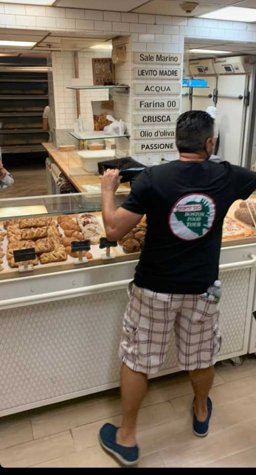 A person in a black shirt and plaid shorts examines baked goods in a bakery display case.