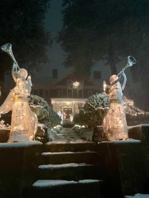 Two illuminated angel figures with trumpets stand at the entrance of a festively decorated house in a snowy night scene.