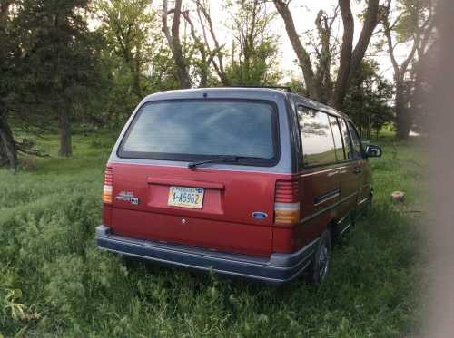 A red van parked in a grassy area surrounded by trees, viewed from the rear.