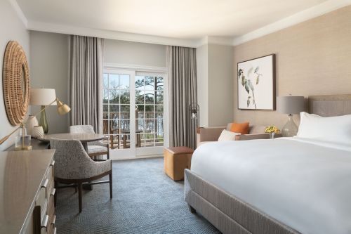 A modern hotel room featuring a large bed, seating area, desk, and a view of the outdoors through large windows.