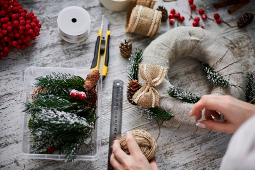 A person crafting a holiday wreath with greenery, pinecones, and decorative elements on a rustic table.