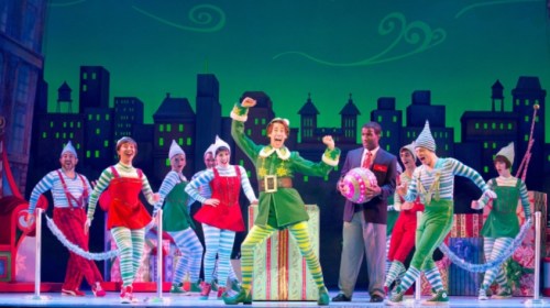 A lively stage performance featuring actors in colorful costumes, celebrating with festive decorations and a city backdrop.