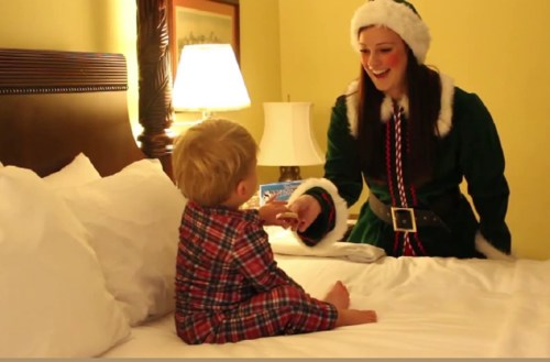 A smiling woman in a festive elf costume interacts with a young boy in pajamas on a hotel bed.