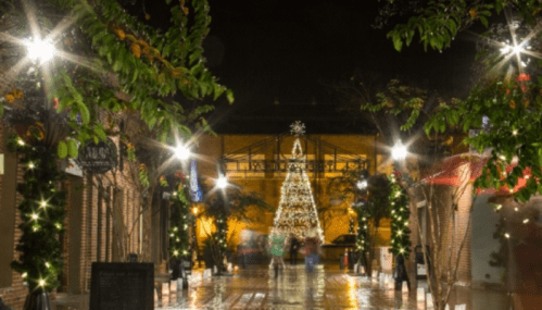 A festive street scene at night, featuring a lit Christmas tree and glowing lights among trees and buildings.