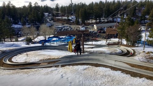 A scenic view of a snow-covered hill with a winding slide and a chairlift, surrounded by trees and a parking area.