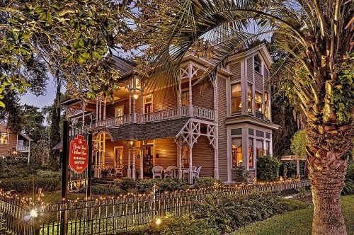 A charming two-story house with a wraparound porch, surrounded by greenery and illuminated by soft lights at dusk.