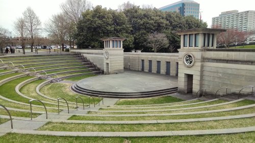 An outdoor amphitheater with stone seating and a stage, surrounded by greenery and trees.