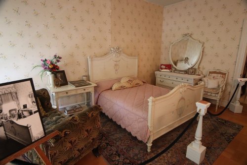 A vintage bedroom featuring a pink quilted bed, ornate mirror, dresser, and floral wallpaper.