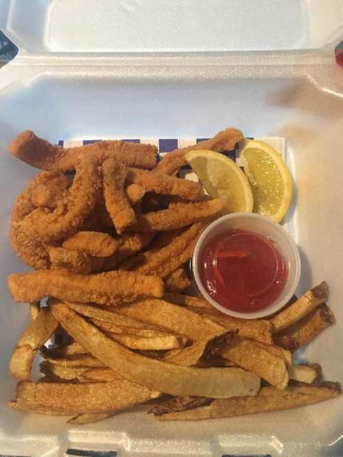 A takeout container with golden fried fish strips, crispy fries, lemon wedges, and a small cup of dipping sauce.
